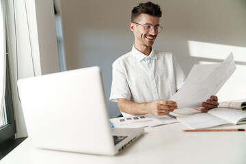 Photo of smiling unshaven man working with laptop and papers at table