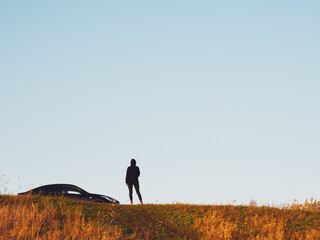 Obraz premium A girl in black clothes stands next to a black sedan car on a hill, against a dim sky, copy space. Concept of auto tourism and travel. Silhouette of a girl and a car at sunset in autumn