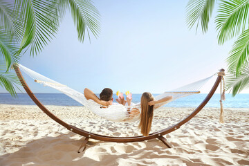 Couple with refreshing cocktails relaxing in hammock under green palm leaves on beach