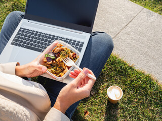 Woman sits on park bench with laptop and take away lunch box, cardboard cup of coffee. Healthy bowl...