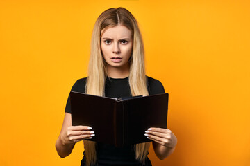 Surprised young woman holding a book on a yellow background. Blonde thoughtful reading a book