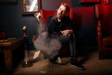 cheerful red-haired guy in a dark room. close-up photo. young man smokes a hookah while sitting in...