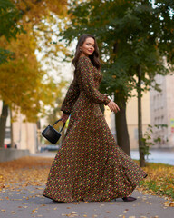 Elegant caucasian brunette woman with long wavy hair in long brown dress with floral print walking...