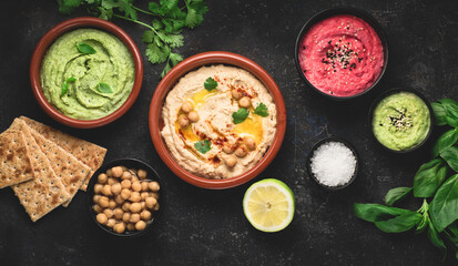 Flat lay of various types of vegetarian hummus based on herbs and beets, basil and coriander on a dark rustic background. Healthy food concept