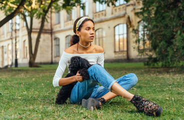 Stylish attractive hispanic woman sitting with a dog on the grass in the park and relaxing on the weekend. Serious lady spends time with a pet in the fresh air.