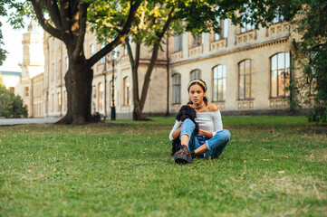 Attractive hispanic woman sitting on the lawn against the backdrop of an old building and a tree with a black little dog in her arms and looking at the camera with a serious face.