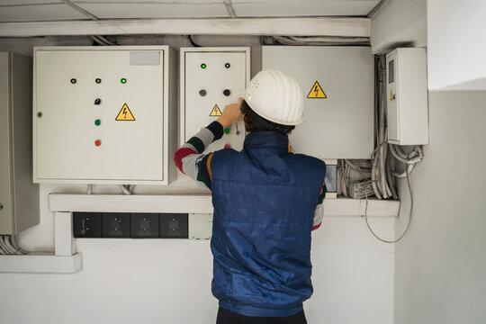 An electrician in an engineering helmet opens the switch box. The technician works in the electrical control room. Service of power distribution switchboards. Back view
