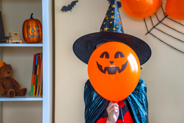 Boy in a black cape and a witch's hat holds a balloon with the image of a pumpkin jack in his face