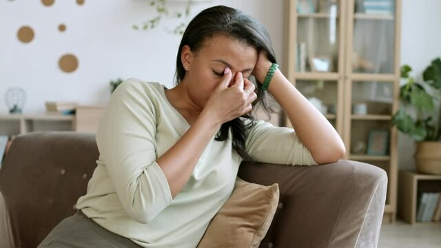 Pan shot of upset and tired mixed-race woman wearing casual clothes sitting on couch at home and touching bridge of her nose