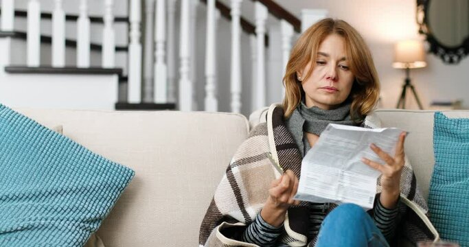 Portrait of Caucasian beautiful middle-aged woman sitting in plaid in cozy room and reading medicine prospectus indoors. Pretty sick female holding pills in hand at home. Treatment concept