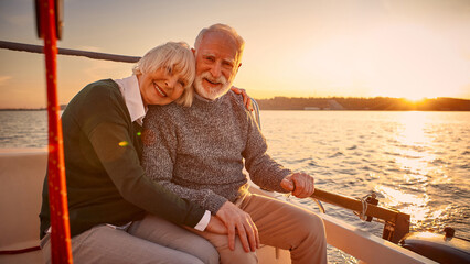 Enjoying amazing sunset. Happy senior couple, elderly man and woman holding hands, hugging and relaxing together while sitting on the side of yacht deck floating in the sea