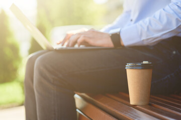 Cropped shot of a male freelancer working online on laptop and drinking coffee while sitting on the bench outdoors on a sunny day, side view
