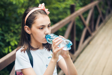 Portrait of a charming child holding a bottle of water, in t-shirt with a backpack on his back, standing in the open air