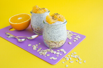 Pudding with chia, oat flakes and orange in the glass jars on the violet cutting board on the yellow background