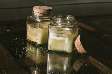 two aromatic ascent candles in a glass square port with cork lead on a glass rustic table top.