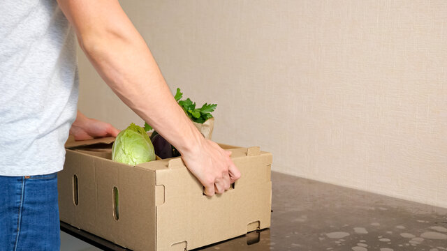 Delivery Service Man Puts Cardboard Box With Vegetables On Grey Table Against White Background Side View Closeup