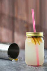Tarro de cristal con tapa dorada y pajita de plástico rosa, con rodajas de cítricos y un refresco amarillo en su interior, sobre un suelo gris y pared de madera con coctelera detrás