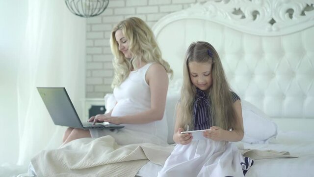 Confident pregnant woman using laptop and admiring cute little girl playing video games online at front. Portrait of happy Caucasian mother surfing Internet while daughter having fun using gadget.