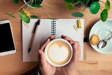 Cup of coffee in woman's hands in the morning with notebook, pen, mobile phone and plants on wooden office desk during the break. Fresh italian cappuccino in cafe. Closeup and flat lay.