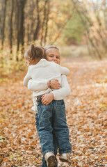 Fototapeta premium Two cheerful small sisters in white christmas sweaters and jeans hugging in the park in warm autumn day