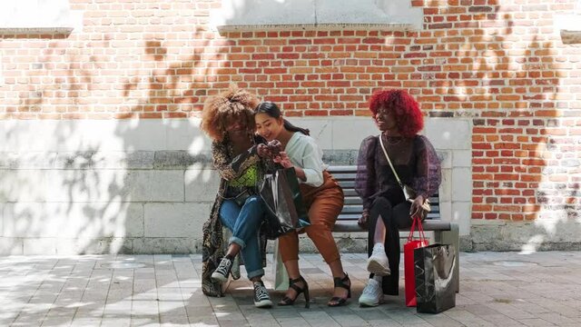 multiracial female friends with shopping bags sitting on a bench and speaking together. Happy smiling generation z girls hang out together.
