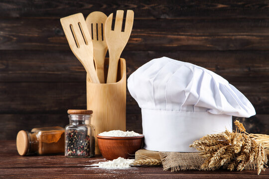 Chef hat with spices in jars, cooking cutlery and bowl with flour on wooden background