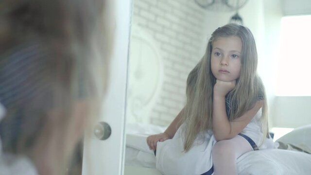 Sad little girl looking in mirror and sighing. Reflection of upset Caucasian brunette child with brown eyes sitting on bed at home and thinking. Unhappy kid alone indoors.