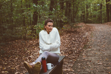 Elegant woman sitting on bench in forest on cold autumn day, she is shivering and trying to get...