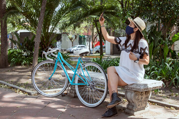 Chica con cubrebocas, tomándose una selfie  en el parque, usando un vestido blanco y un sombrero con una bicicleta al lado.