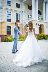 Naklejka premium Young stylish guy in a suit the groom and the bride beautiful girl in a white dress with a train walk in the park on their wedding day