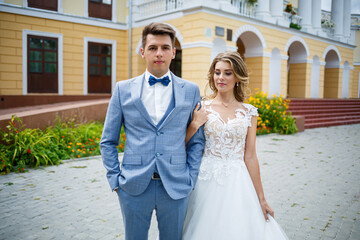 Naklejka premium Young stylish guy in a suit the groom and the bride beautiful girl in a white dress with a train walk in the park on their wedding day