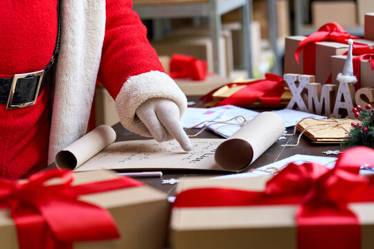 Santa Claus wearing costume reading parchment roll letter wish list standing at table with presents in workshop preparing for holiday on xmas eve. Merry Christmas wishlist background concept, closeup.