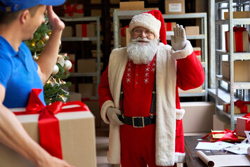 Happy old bearded Santa Claus waving hand to courier delivering Christmas present parcel on xmas eve standing in workshop. Male deliveryman holding gift box, fast express shipping delivery concept. - obrazy, fototapety, plakaty