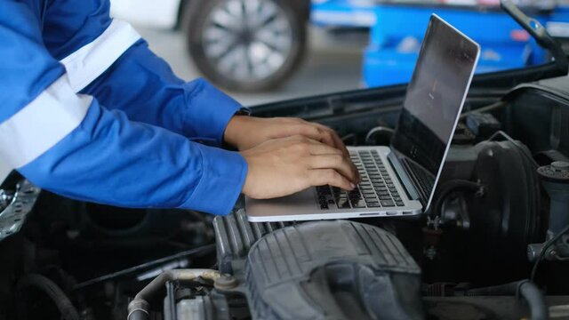Mechanic Asian man using laptop computer examining tuning fixing repairing car engine automobile vehicle parts using tools equipment in workshop garage support and service in overall work uniform
