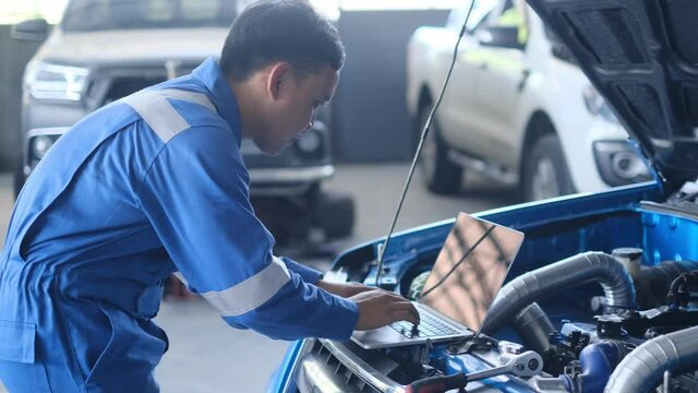 Mechanic Asian man using laptop computer examining tuning fixing repairing car engine automobile vehicle parts using tools equipment in workshop garage support and service in overall work uniform