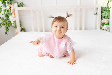 Fototapeta premium cheerful baby girl six months old sitting in a bright beautiful room on a white bed in pink clothes and smiling