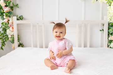 cheerful baby girl six months old sitting in a bright beautiful room on a white bed in pink clothes and smiling