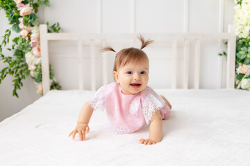 Fototapeta premium cheerful baby girl six months crawling in a bright beautiful room on a white bed in pink clothes and smiling