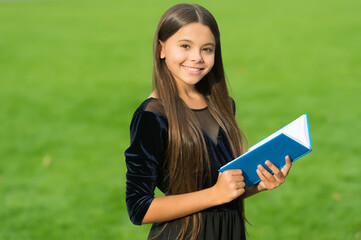 Fototapeta premium Happy small kid in school uniform smile holding study book green grass sunny summer outdoors, education