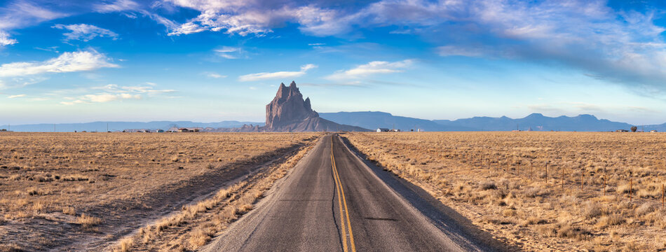 Panoramic View of a road in a dry desert with a Shiprock mountain peak in the background. Sunny and Cloudy Morning Sunrise Artistic Render. Taken at Rattlesnake, New Mexico, United States.