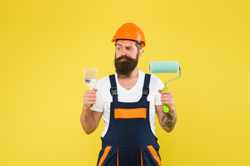 Skilled painter with serious look in working overalls hold paint brush and roller for painting work...