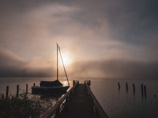 Obraz premium wooden jetty with a sailboat in the water of a lake in fog at sunrise