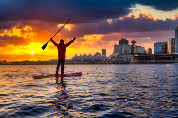 Fototapeta premium Adventurous man is paddle boarding near Downtown City during a vibrant winter sunrise. Dramatic Sky Artistic Render. Taken in Coal Harbour, Vancouver, British Columbia, Canada. Colorful Sky Overlay