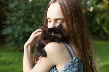 Naklejka na ściany i meble Black fluffy kitten cat in the arms of a beautiful young girl with long hair in summer. Outside.
