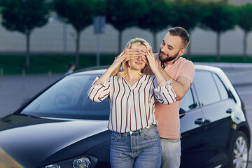 Man giving surprise to woman by purchasing a new car. Young couple buys a car, man and woman are standing near the car on the street. Surprise gift for a loved one