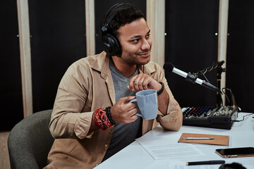 Portrait of young male radio host going live on air, talking with guest, holding a cup of coffee or...