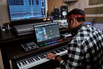 Young man, male artist looking focused while playing keyboard synthesizer, sitting in recording studio