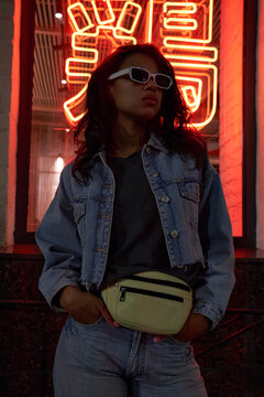Cool African American hipster gen z lady wears trendy glasses denim clothes standing under neon night sign. Stylish black young fashion model illuminated in 80s 90s retro style. Translation: Noodles