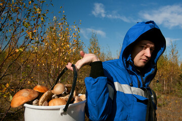 Man in blue protective clothing with bucket of mushrooms in the north autumn forest. Sunny october day