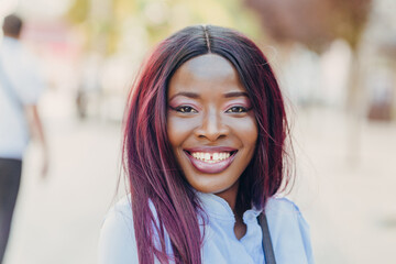 a smiling young African American girl with pink hair and a blue shirt walking outside on a sunny day.